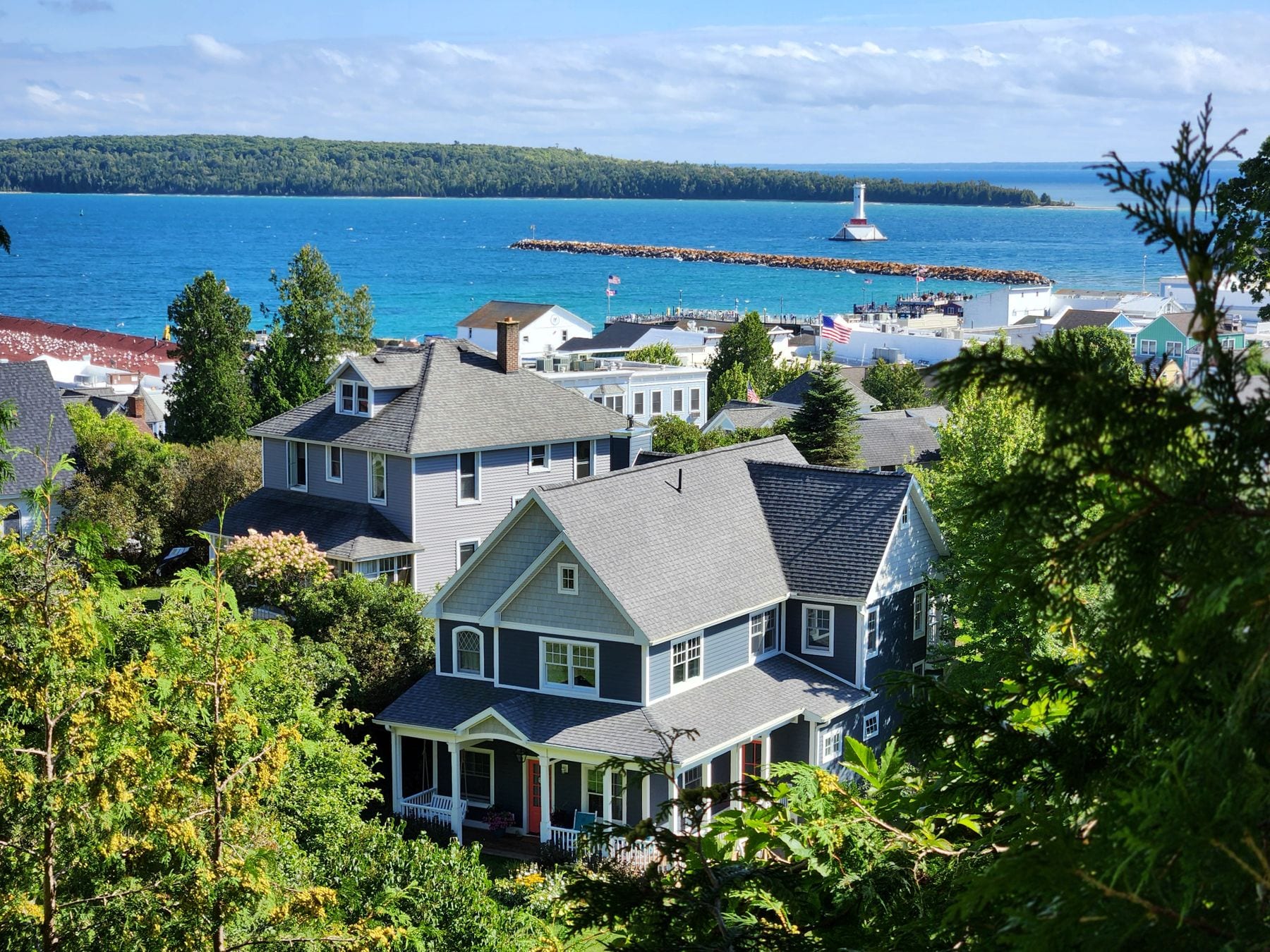 Waterfront homes on a wooded hillside above a Northern Michigan harbor, with a lighthouse and breakwater in the distance